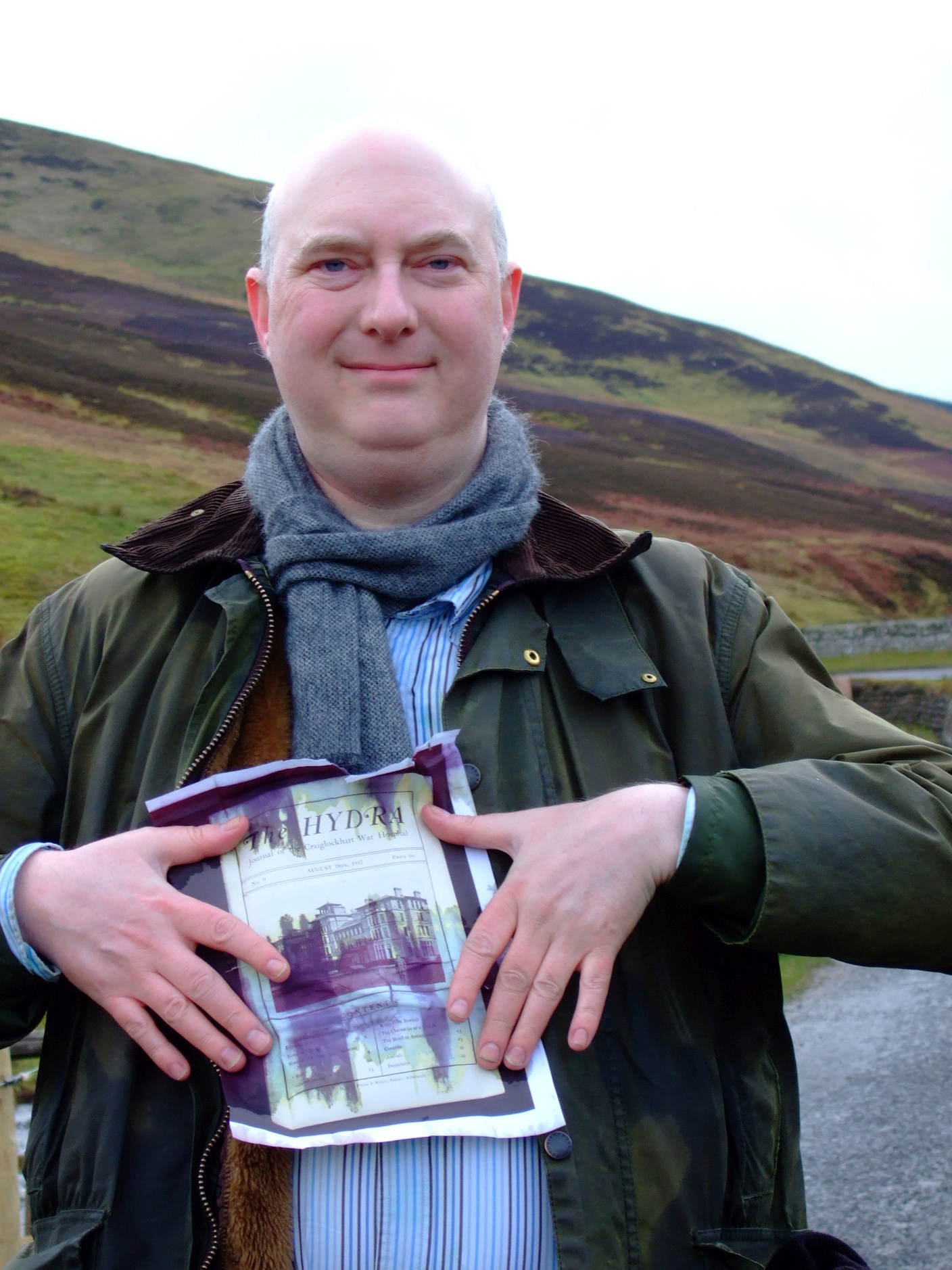 A bald man in a Barbour jacket holds a soaked photocopy of a magazine to his chest with the Pentland hills rising behind.