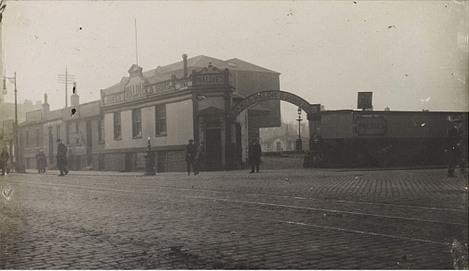 A fogged image of the arched corner entrance to a coal merchant. People wander the cobbled street which has tram tracks visible