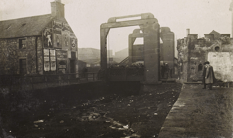 A metal swing bridge is visible above a drained canal basin full of litter. Onlookers survey the scene. A nearby building is covered in advertising posters.