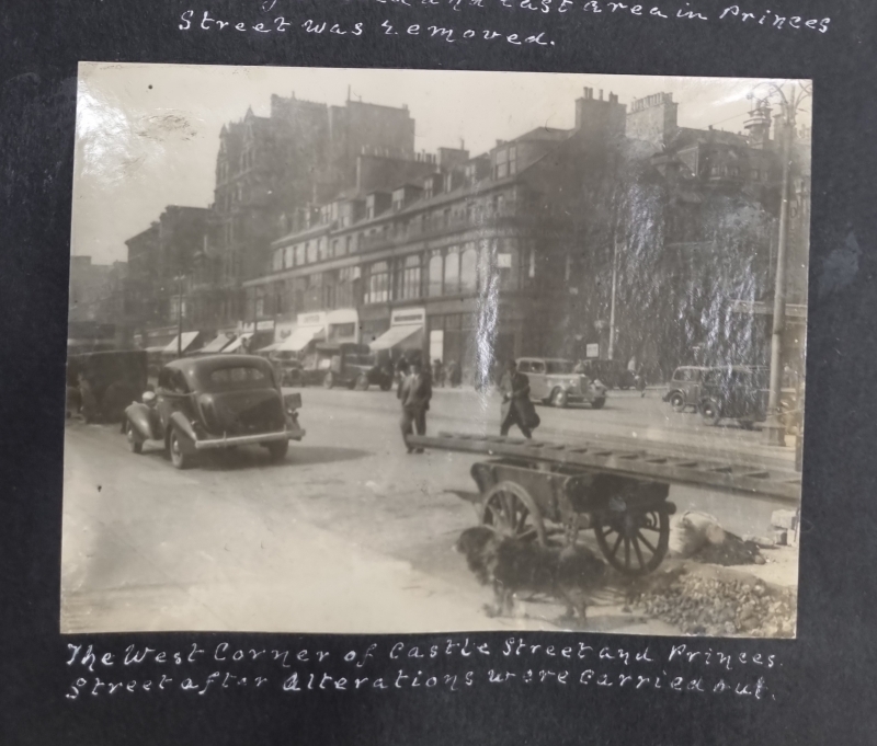 A fogged and faded 1938 city street scene with heavy traffic and pedestrians crossing in front of city centre shops.