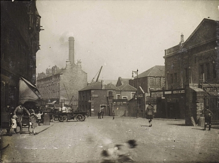 A busy city street: a man in a kilt walks away from us, a woman to the right struggles with children, and in the distance a tall industrial chimney smokes.